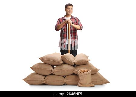 Portrait complet d'un fermier mâle avec une pelle debout derrière des sacs de burlap avec des pommes de terre isolées sur un fond blanc Banque D'Images