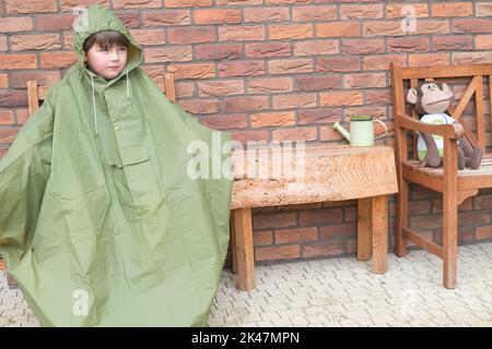 Garçon dans le manteau de pluie vert est assis et en attente de l'arrêt de pluie. Automne saison pluvieuse. Arrière-plan du mur de briques. Chaise en bois et conte. Enfant veut Banque D'Images