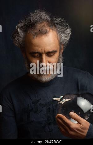 un homme coloré avec des boucles une barbe et une moustache en argent tient une boule de verre transparente dans sa main et regarde vers le bas avec sa tête pendue Banque D'Images