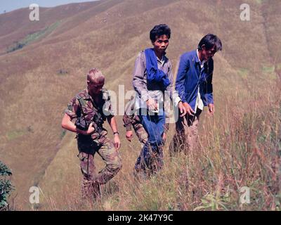 Deux hommes immigrants illégaux ('IIS'), juste arrêtés à Hong Kong, sont escortés jusqu'à Robin's Nest, une colline près de la frontière dans les nouveaux Territoires de Hong Kong, par deux soldats du régiment de Cheshire, armée britannique. Oct 1984. Le soldat à gauche porte un vieux kit radio. Les immigrants illégaux portent les vêtements de travail en coton bleu typiques de l'époque. Banque D'Images