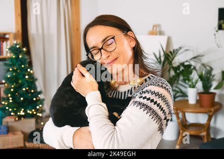 Jeune femme souriante embrassant chat domestique noir dans des lumières de Noël festives dans une pièce moderne avec des cadeaux et la décoration de noël. Joyeuses vacances d'hiver Banque D'Images
