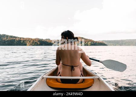 fille caucasienne portant un maillot de bain assis à l'intérieur d'un kayak sur son dos et regardant le côté pagayer sur le lac vers l'île, le lac tarawera Banque D'Images