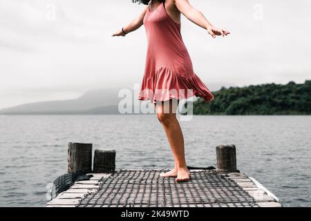 jeune femme caucasienne vêtue d'une robe rose avec ses bras tendus dansant pieds nus sur l'ancienne jetée en bois du lac calme, le lac tarawera, nouvelle-zélande Banque D'Images