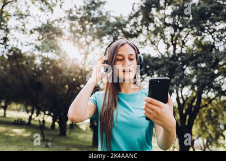 Une adolescente souriante, portant un t-shirt turquoise et un casque, fait un appel vidéo avec son mobile dans un espace naturel Banque D'Images