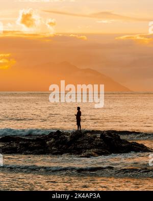 Une photo verticale d'un magnifique coucher de soleil en mer avec la silhouette d'un homme debout sur des rochers sous un ciel doré Banque D'Images