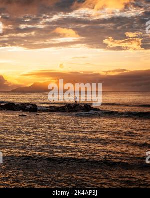 Une photo verticale d'un magnifique coucher de soleil en mer avec la silhouette d'un homme debout sur des rochers sous un ciel doré Banque D'Images