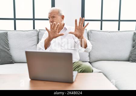 Homme âgé utilisant un ordinateur portable à la maison assis sur le canapé peur et terrifié avec l'expression de peur stop geste avec les mains, criant dans le choc Banque D'Images