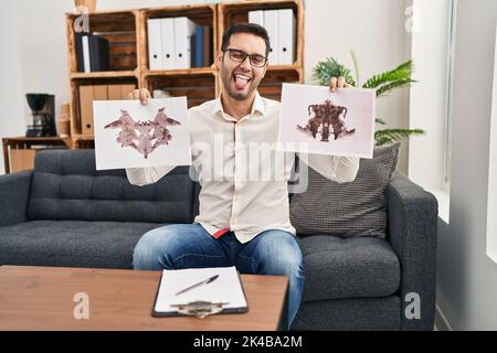 Jeune homme hispanique avec la barbe tenant le test de rorschach collant la langue dehors heureux avec l'expression drôle. Banque D'Images