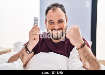 Homme hispanique de grande taille avec barbe dans le lit tenant des pilules criant fier, célébrant la victoire et le succès très excité avec les bras levés Banque D'Images