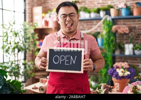 Jeune homme chinois travaillant au fleuriste tenant le signe ouvert collant la langue dehors heureux avec l'expression drôle. Banque D'Images