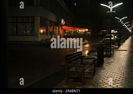 Pluvieux tard dans la soirée dans le parc, une rangée de bancs en bois, des reflets au néon des lampes de rue dans l'asphalte humide. Banque D'Images