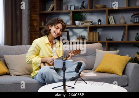 Blogueur avec téléphone mobile et microphone. Une belle hipster fille dans une chemise jaune joue et chante sur le ukulele sur le canapé dans l'appartement Banque D'Images