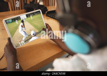 Un garçon afro-américain porte des écouteurs à la maison pour regarder un match de baseball sur tablette. Sports, compétition, divertissement et technologie concept numérique co Banque D'Images