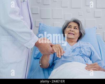 Sourire heureux belle asiatique vieille femme patiente en robe bleu clair couché sur le lit tandis que le médecin masculin en costume blanc tenant sa main et donnant l'intérieur Banque D'Images