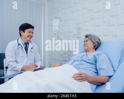 Souriant asiatique médecin en costume blanc prendre des notes tout en discutant heureux asiatique âgée, femme âgée qui s'est couché sur le lit avec la solution saline Banque D'Images