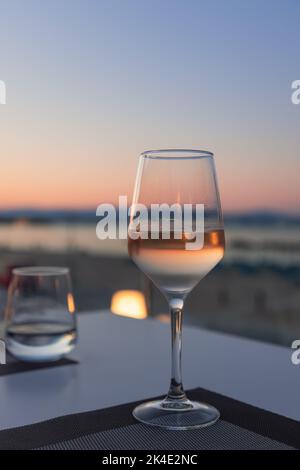 La lueur rose d'un coucher de soleil avec la mer et la plage de sable se reflète dans un verre concentré de vin blanc sur une table dans un café de plage Banque D'Images