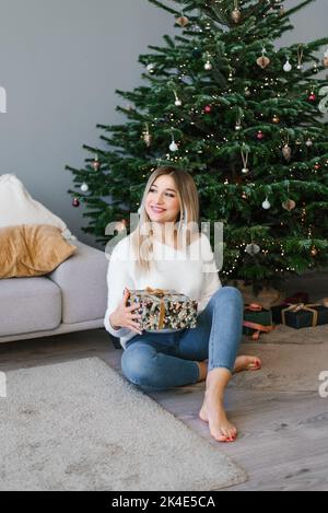 Belle femme est assise sur le sol près de l'arbre de Noël dans la salle de séjour et tient un cadeau surprise dans ses mains et souriant Banque D'Images