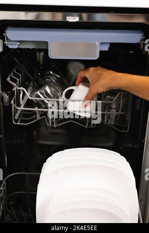 Gros plan sur la main d'une femme qui charge des plats, les vide ou les déchargeant d'un lave-vaisselle automatique ouvert intégré avec des plats à l'intérieur de la cuisine. Banque D'Images