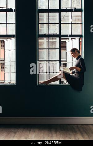 Vue latérale d'une femme asiatique assise sur le rebord de la fenêtre et lisant un livre. Photo verticale d'une femme lisant un magazine près de sa fenêtre de bureau. Banque D'Images