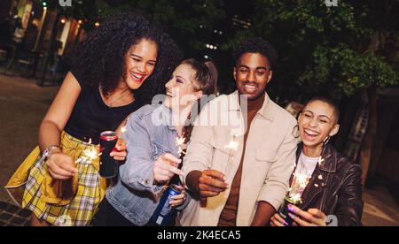 Des amis de la ville célèbrent la saison des fêtes avec des fêteurs. Groupe de jeunes heureux tenant des lumières du bengale et des canettes de bière la nuit. Amis joyeux Banque D'Images