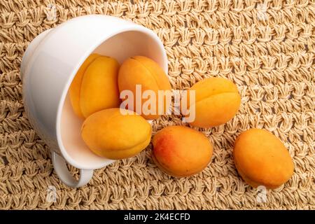 Plusieurs abricots jaunes d'ananas dans une tasse blanche, gros plan, sur un tapis de paille, vue de dessus. Banque D'Images