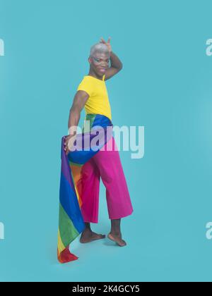 Homme afro-américain plein de corps heureux avec le drapeau LGBT enveloppé autour des hanches touchant la tête et regardant la caméra avec le sourire sur fond turquoise Banque D'Images