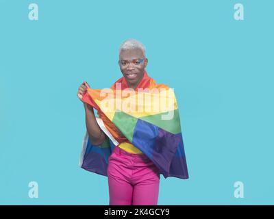 Modèle africain américain positif avec maquillage regardant la caméra avec le sourire et enveloppant le drapeau arc-en-ciel autour des épaules tout en soutenant les movemen LGBT Banque D'Images