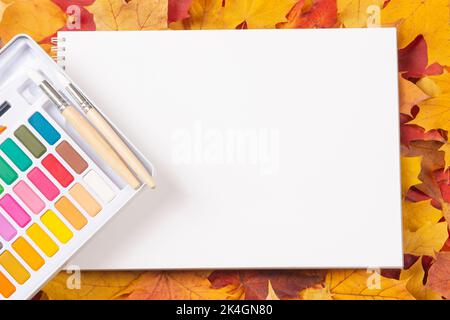 Composition d'automne avec carnet à croquis ouvert vierge et peinture aquarelle sur fond rouge, orange et jaune feuilles d'automne d'érable d'automne. Maquette, vue de dessus Banque D'Images