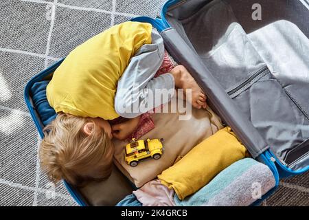 Un enfant triste se trouve dans une valise qui attend le départ et joue avec une voiture-jouet. Déménagement de la famille, vol du pays Banque D'Images