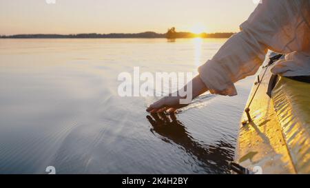 La personne navigue sur le kayak touchant l'eau avec la main faisant de petites vagues de gros plan. Anonyme aime le canoë et le tourisme. Le soleil se lève à l'horizon Banque D'Images