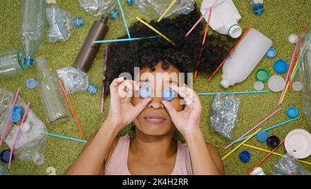 Une femme afro-américaine met les bouchons en plastique des bouteilles sur les yeux. La femme au foyer se trouve sur un tapis près des ordures éparpillées, ce qui met l'accent sur le problème de gros plan Banque D'Images