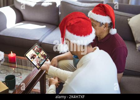 Père biracial et fils adulte dans les chapeaux de santa faire tablette de noël appel vidéo de groupe avec des amis. noël, festivité et technologie de communication. Banque D'Images