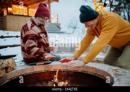 Couple senior assis et chauffant ensemble au foyer extérieur dans leur jardin d'hiver. Banque D'Images