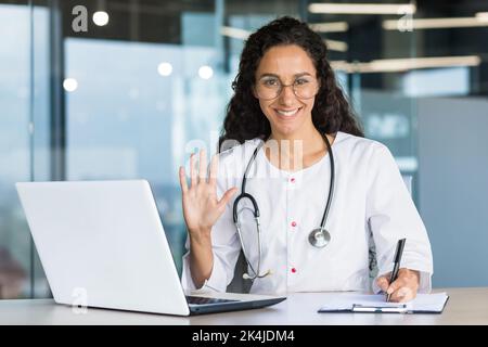 Portrait d'une femme hispanique médecin vétérinaire. Il est assis au bureau, mène une consultation en ligne avec un ordinateur portable, regarde et fait des vagues à la caméra. Enregistre le diagnostic, le traitement de l'animal. Banque D'Images