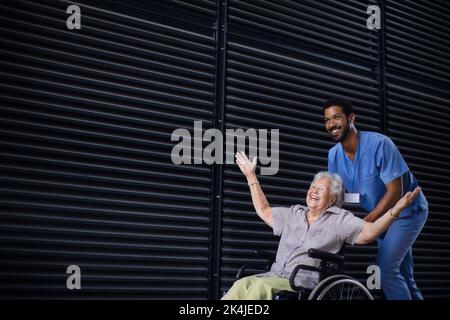 Homme soignant poussant une femme âgée excitée en fauteuil roulant dans la rue, en appréciant le temps ensemble. Banque D'Images
