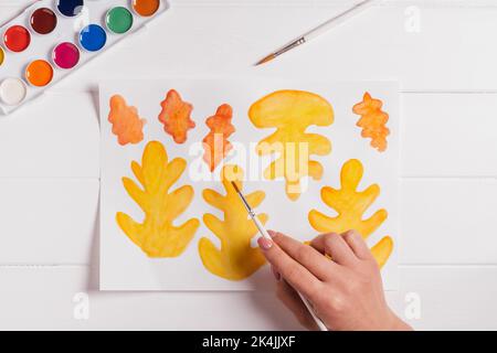 Femelle dessin à la main de feuilles d'automne de différentes formes, tailles et couleurs avec aquarelles et pinceau sur table blanche en bois. Vue de dessus. Bricoler à la main Banque D'Images