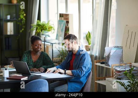 Un jeune homme d'affaires heureux pointant ses notes dans un livre publicitaire lors de la discussion de données avec une femme africaine américaine collègue Banque D'Images