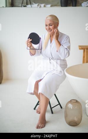 Joyeuse femme bien entretenue examinant sa peau dans le miroir à main Banque D'Images