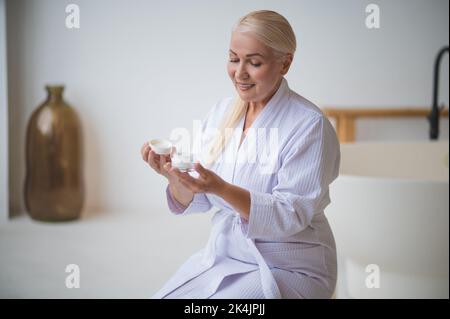 Joyeux femme assise dans la salle de bains examinant le produit de soin de la peau Banque D'Images