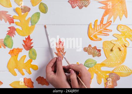 Femelle dessin à la main de feuilles d'automne de différentes formes, tailles et couleurs avec aquarelles et pinceau sur table blanche en bois. Vue de dessus. Bricoler à la main Banque D'Images