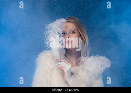 femme blonde avec maquillage d'hiver dans la veste en fausse fourrure tenant la plume blanche sous la neige tombant sur bleu foncé, image de stock Banque D'Images