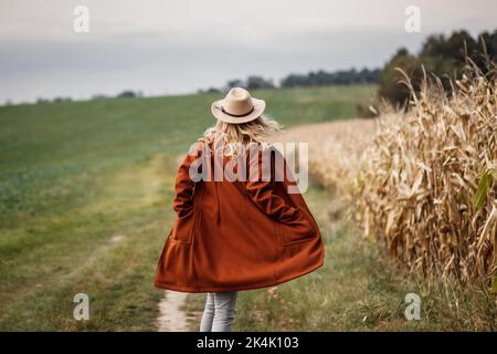 Bonne femme avec chapeau de cowboy et manteau rouge courant au champ de maïs. Saison d'automne en campagne. Collection automne Banque D'Images