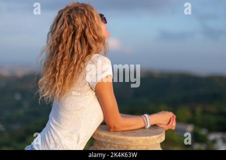 Jeune femme caucasienne positive en lunettes de soleil appréciant le coucher de soleil à l'extérieur, vue arrière Banque D'Images