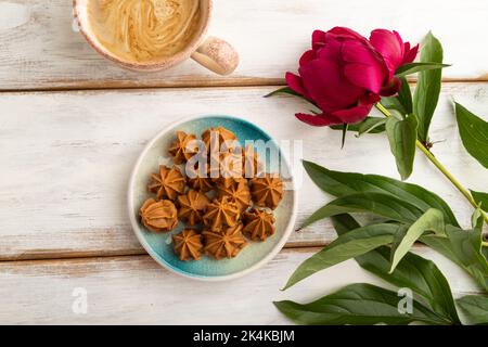 Bonbons au caramel doux faits maison sur une assiette bleue et une tasse de café sur fond de bois blanc, décoration de fleurs de pivoine. Vue sur le dessus, plat, gros plan. Banque D'Images