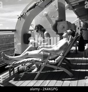 Fin des années 1960, historique, croisière heureuse, un couple adulte en maillot de bain se détendant dans des transats sur le pont du navire de croisière Norweigian-America, MS Sagafjord. Le passager est couché en train de prendre du soleil, tandis que la dame lit un livre. Banque D'Images