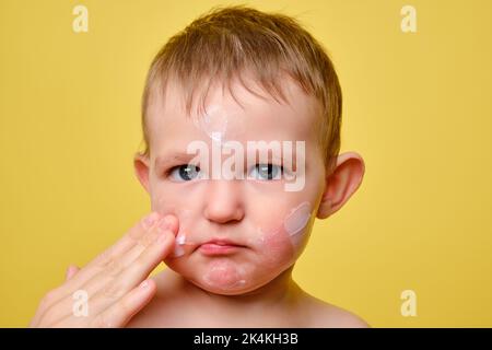 La mère sent la crème allergique sur le visage du bébé tout-petit, fond jaune studio. Portrait en gros plan d'un bébé mignon avec de la crème sur ses joues. Enfant âgé d'un an Banque D'Images