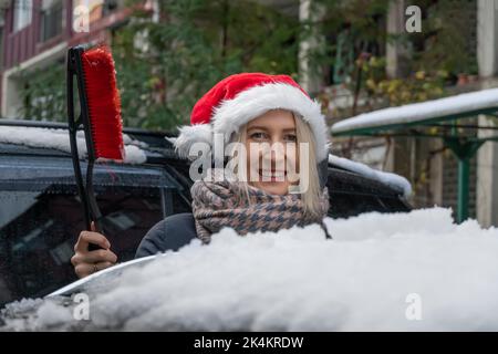 Une jeune femme dans un chapeau de Père Noël avec une brosse à neige est debout près de la voiture, va nettoyer sa voiture Banque D'Images