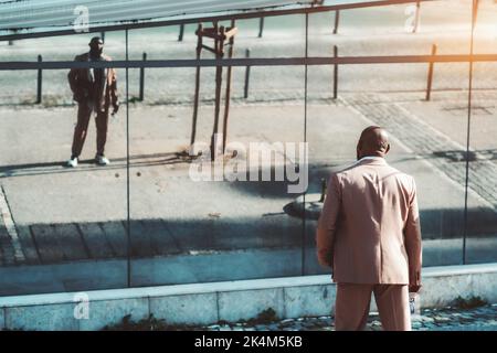 Vue de l'arrière d'un élégant homme noir bald dans un costume taillé rose qui donne sur une façade murale miroir d'un immeuble de rue qui le reflète entièrement Banque D'Images