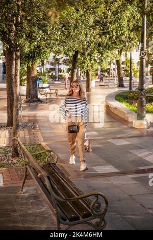 Belle femme chien de marche dans la ville. Bonne femme portant des lunettes de soleil et des vêtements décontractés tout en emportant son chien pour une promenade. Banque D'Images