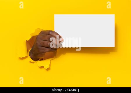 Homme noir tenant une feuille blanche vierge à travers le trou sur fond de papier jaune, maquette Banque D'Images